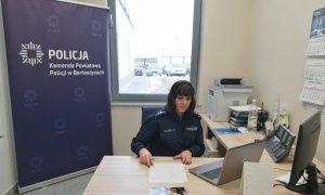 policjantka w czasie lekcji online  siedzi przed ustawionym na biurku laptopie, za nią baner z zapisem Komenda Powiatowa Policji w Bartoszycach